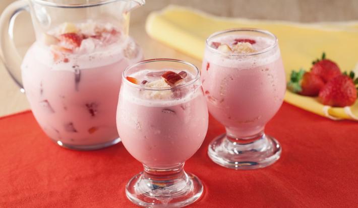 Fotografia em tons de vermelho em uma mesa de madeira com uma toalha vermelha, um pano amarelo com três morangos em cima. Ao centro, dois copos de vidro com a sangria de Leite Moça com morangos e atrás uma jarra de vidro com a sangria.