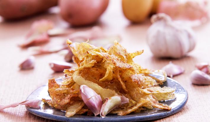 Foto da receita de casca de batata no alho, servida em um prato azul, decoradas com dentes de alho, numa mesa clara com mais batatas e alhos inteiros ao fundo