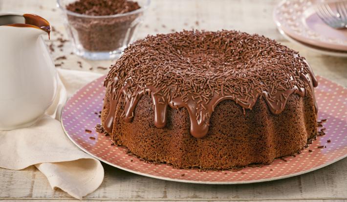 Imagem de um bolo redondo de chocolate com cobertura de brigadeiro e chocolate granulado sobre um prato rosa com bolinhas brancas.
