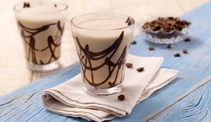 Foto da receita de smoothie de cappuccino e coco. Observa-se dois copos transparentes com calda de chocolate e a bebida, sobre um guardanapo bege em uma superfície de madeira metade bege e metade azul clara. Como decoração, grãos de café no fundo