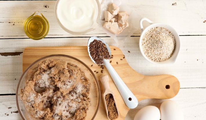 Fotografia em tons de branco e marrom de uma bancada branca com uma tábua de madeira clara, sobre ela um recipiente de vidro com ingredientes. Ao lado dois ovos, e recipientes com azeite, iogurte, fermento e aveia.