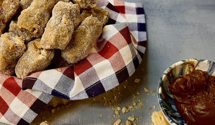 Foto da receita de paglia de doce de leite servida em diversas porções em uma cesta com um pano quadriculado vermelho e preto sobre uma mesa cinza, com um bowl de porcelana com doce de leite ao lado.
