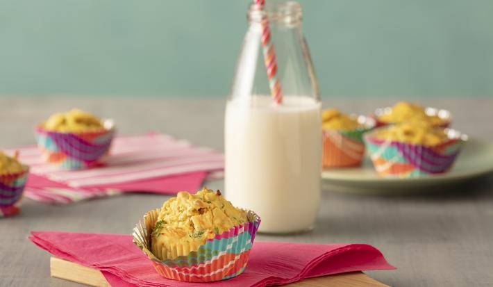 Fotografia em tons de rosa, em uma bancada de madeira de cor cinza. Ao centro, uma tábua de madeira com um pano rosa em cima. Em cima do pano está o muffin e ao lado há um pote de vidro contendo leite. AO fundo alguns muffins espalhados.