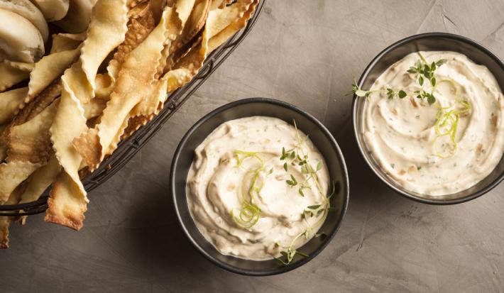fotografia em tons de cinza, branco e pardo de uma bancada cinza vista de cima. Contém dois potinhos redondos e pretos com uma pasta de creme, e ao lado biscoitinhos para servir junto ao creme.