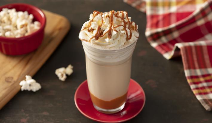Foto de uma bancada de madeira, com um pires vermelho no centro e sobre ele um copo com a bebida em camadas marrom, branca, decorada com chantilly, pipocas e doce de leite. Há um pano vermelho xadrez, uma tábua e um pote com pipocas ao fundo