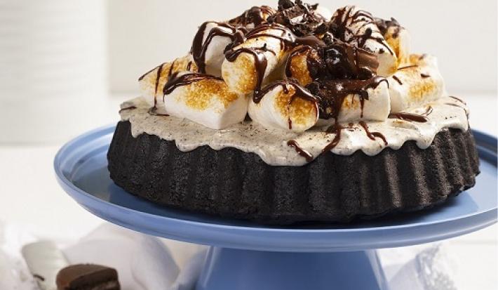 Fotografia em tons de branco, em uma bancada branca de madeira. Ao centro, um boleiro azul contendo a torta s'more. Ao lado, uma faca e ao fundo um vaso branco de flores amarelas.