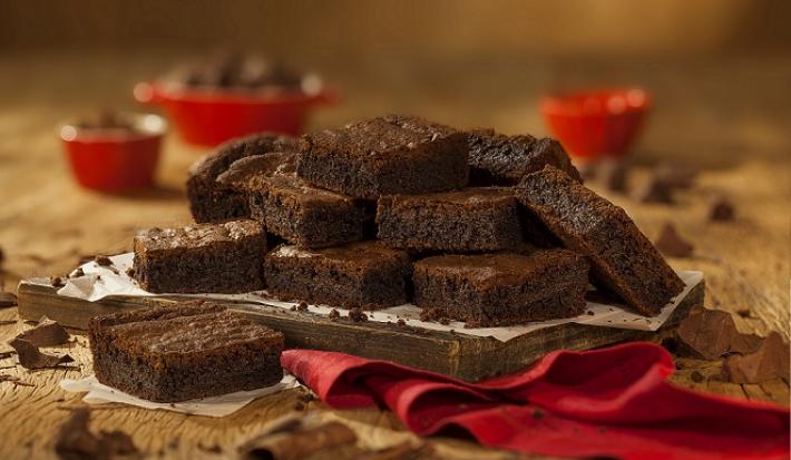 Fotografia em tons de marrom em uma bancada de madeira de cor marrom. Ao centro um pano vermelho contendo uma tábua de madeira com os brownies em cima. Ao redor, há vários pedaços de chocolate espalhados.