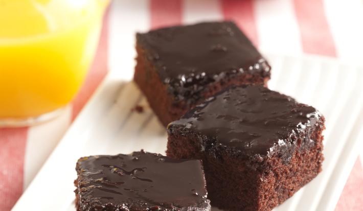 Foto com 3 pedaços quadrados de bolo de Nescau e cobertura de Nescau. No fundo da foto, há uma jarra com suco de laranja e 2 maças em um prato vermelho