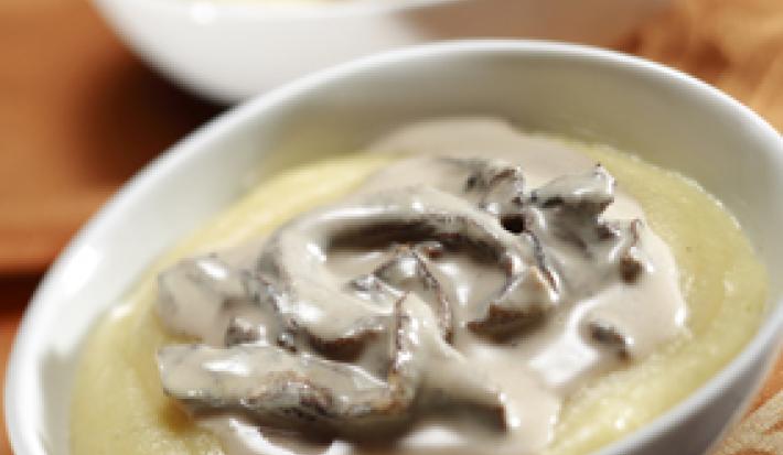Fotografia em tons de amarelo em uma bancada de madeira, um pano bege e dois recipientes ovais brancos com a polenta coberta com molho de funghi.