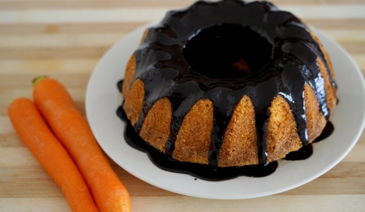 Bolo de cenoura inteiro, assado em forma de buraco no meio, está sobre um prato branco. Ele está coberto com uma generosa calda de chocolate escorrida pelas laterais. Ao lado do prato, há duas cenouras cruas, inteiras..