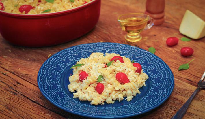 Imagem de um prato azul com macarrão com tomate e queijo feta e ao lado, um pedaço de queijo, tomatinhos e azeite.