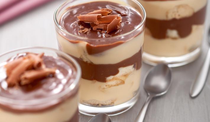 Fotografia em tons de marrom em uma bancada de madeira clara com um pano rosa de fundo e três taças de vidro com pavê de chocolate com maracujá e raspas de chocolate para enfeitar.