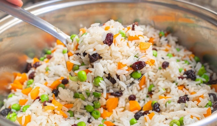 Dentro de uma panela, há arroz branco com uvas-passas, cenouras e ervilhas.