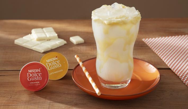Foto da receita de mocha branco com latte macchiato servida em um copo de vidro alto sobre um pratinha laranja em cima de uma mesa de madeira com uma barra de galak e um paninho ao fundo