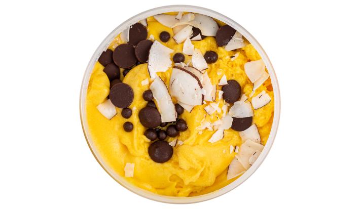 Fotografia vista de cima de um flan de maracujá, leite MOÇA e leite de coco, e por cima, gotinhas de chocolate e chips de coco. O creme está dentro de um recipiente de vidro na cor branco, sobre um fundo branco.