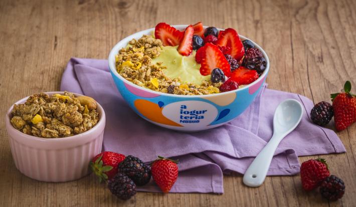 Fotografia em tons de lilás em uma bancada de madeira clara, um bowl colorido com o iogurte de abacate com frutas vermelhas e granola decorando. Ao lado, um potinho com granola e frutas espalhadas na bancada.