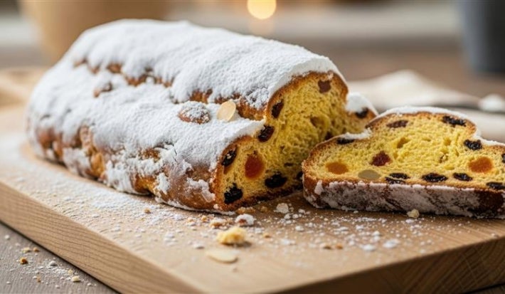 Fotografia de um bolo rrecém-assado, colocado em uma tábua de madeira. O bolo, coberto com açúcar de confeiteiro, revela um interior dourado com frutas secas e amêndoas.