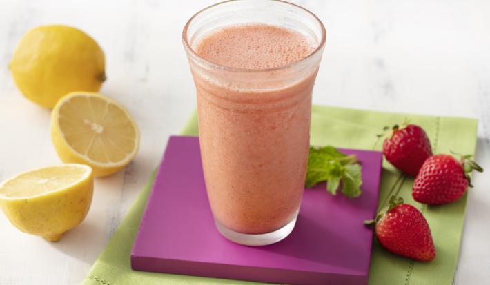 Fotografia em tons de branco, roxo e verde de uma bancada de madeira branca e um paninho verde, sore ele um apoio de copo quadrado roxo com um copo de vidro com o suco de morango. Ao lado três morangos e dois limões.
