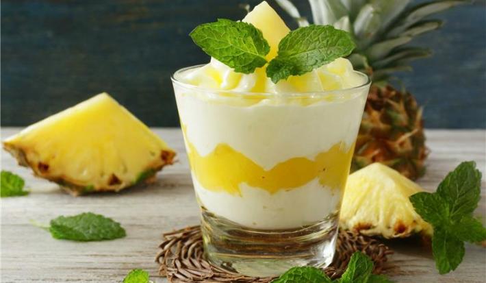 Na imagem consta uma sobremesa de abacaxi no copo montada em camadas. Abaixo temos odoce de abacaxi, coberto por creme branco leve e decorado com pedaços de abacaxi e hortelã