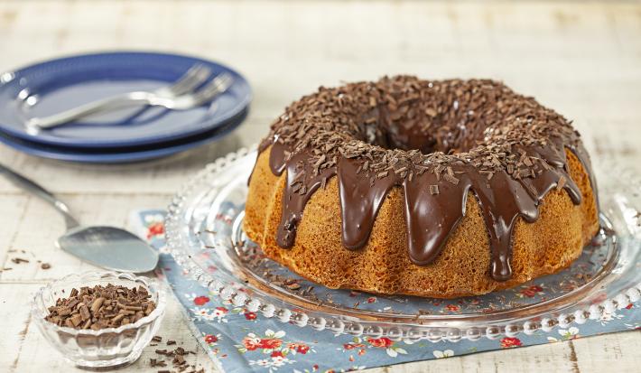 Imagem de um bolo de cenoura com cobertura de brigadeiro e raspas de chocolate sobre um prato redondo transparente.