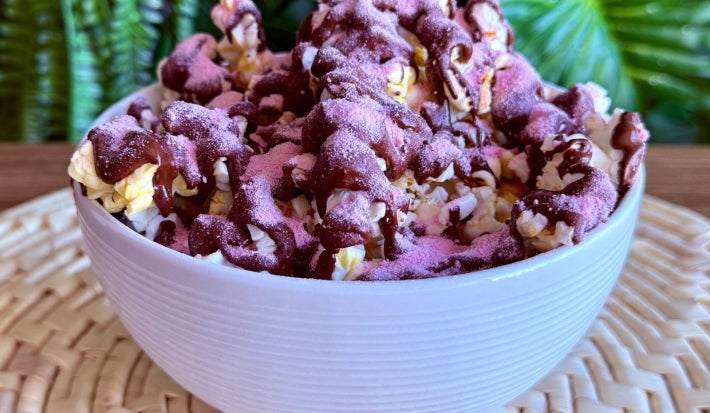 Fotografia em tons de rosa e marrom com um pote branco ao centro. Dentro do prato existe uma receita de pipoca doce coberta com chocolate meio amargo e polvilhada com Nesquik
