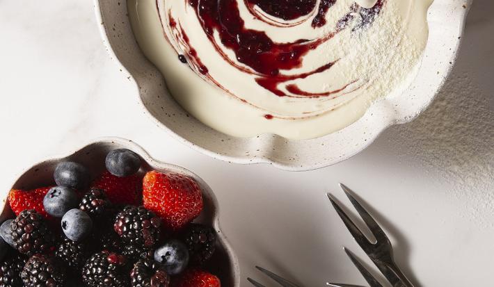 Fotografia em tons de branco, em uma mesa de madeira de cor branca. Ao centro, um recipiente contendo o fondue e ao lado um outro recipiente contendo as frutas. Ao fundo, alguns garfos.