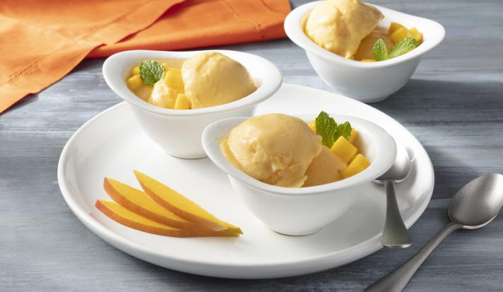 Imagem aproximada da receita de Sorvete Zero Lactose em tom amarelo claro, servidos em três porções em potes brancos, decorados com cubos de manga e folhas de hortelã. A bancada em que estão está decorada com mangas cortadas, um tecido laranja e talheres