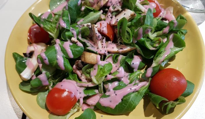 Fotografia vista de cima de uma salada de alface, cenoura, tomates cereja, pepino e beterraba com molho à base de iogurte e ketchup. A salada está em um prato amarelo que está sobre uma mesa branca.