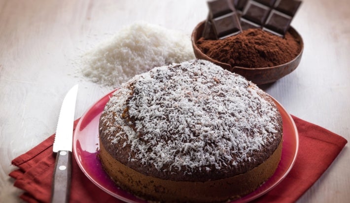Fotografia de um bolo de chocolate em um prato vermelho, coberto com coco ralado. Ao lado, uma faca serrilhada sobre um guardanapo vermelho, com ingredientes como coco e cacau ao fundo.