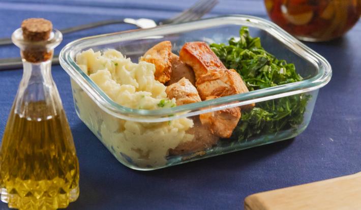 Fotografia mostra uma marmita em pote de vidro com uma porção cremosa de purê de batata-doce no lado esquerdo, cubos de frango grelhado dourados ao meio e couve refogada brilhante e bem verde no canto inferior direito.