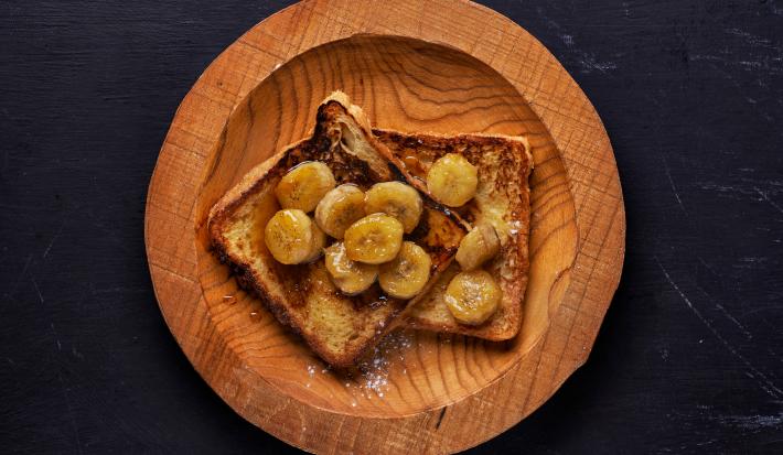 Fotografia em tons de madeira com um prato de madeira ao centro. Em cima do prato existe dois pães de forma cobertos com banana caramelizada e mel