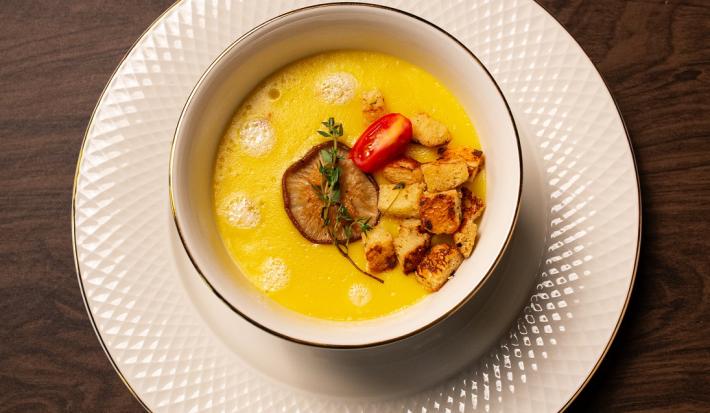 Fotografia em tons de marrom com um prato de sopa branco ao centro. Dentro do prato existe um creme de cor amarela feito com mandioquinhas acompanhado com croutons, cogumelos e tomate cereja.