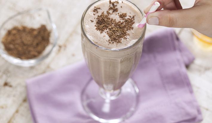 Fotografia em tons e branco e roxo de uma bancada branca com um paninho roxo, sobre ele uma taça de vidro com milk shake e um canudo. Ao lado um recipiente de vidro com raspas de chocolate.