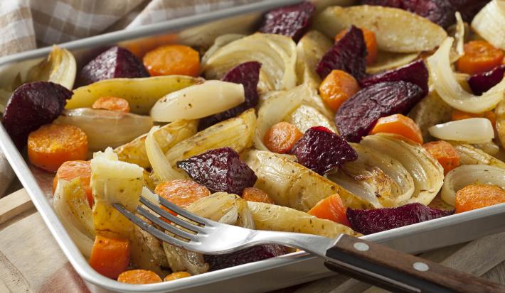 Fotografia em tons de amarelo e roxo, embaixo um pano quadriculado branco e bege, sobre ele uma assadeira de inox com batatas, cenouras, cebolas e beterrabas, há um garfo com cabo de madeira espetando uma batata.