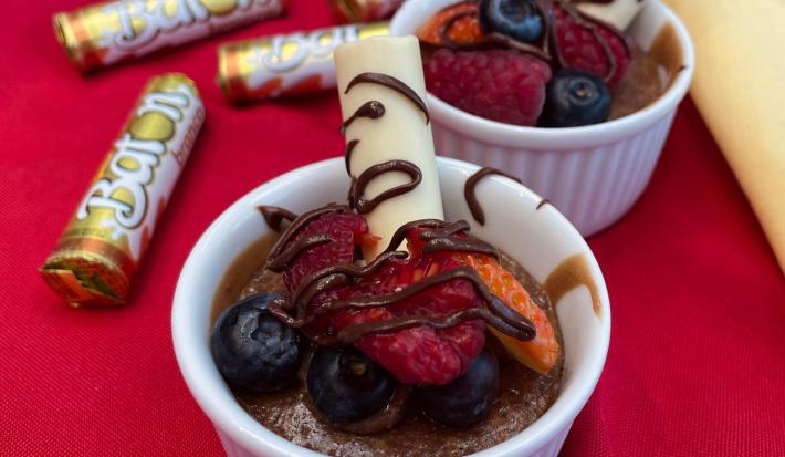 Fotografia em tons de vermelho com dois refratários ao centro. Dentro de cada um deles existe um bolo de chocolate cremoso coberto com morangos, calda de chocolate e BATON branco. Ao fundo existe embalagens de BATON branco.
