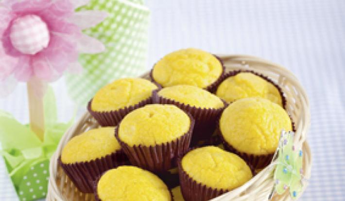 Fotografia em tons de azul, marrom e amarelo, com cestinhas brancas sobrepostas contendo bolinhos em forminhas marrons, sobre toalha quadriculada em branco e azul, com decoração de motivo floral ao lado e fundo em tons de azul.