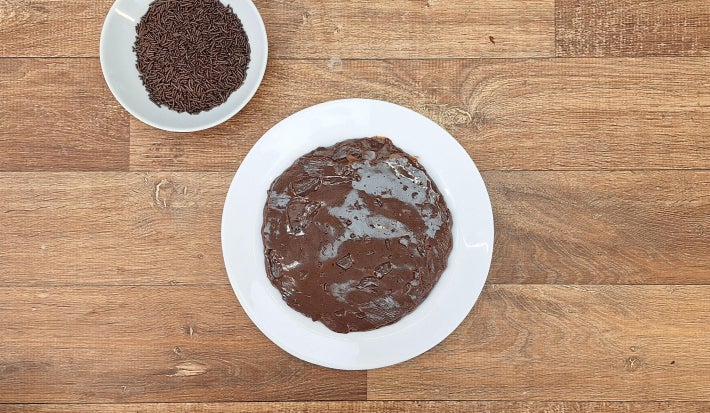 Duas tigelas brancas sobre bancada de madeira. Uma das tigelas tem brigadeiro e a menor tem granulados de chocolate.