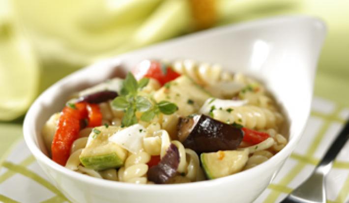 Fotografia em tons de verde em uma mesa de madeira verde com um guardanapo de papel quadriculado em branco e verde e um potinho branco com a salada mediterrânea dentro.