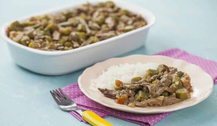 Fotografia em tons de azul e rosa em uma bancada de madeira azul, um guardanapo de pano rosa com bolinhas brancas, um refratário branco retangular com o picadinho de carne com quiabo. Ao lado, um prato rosa redondo com uma porção do picadinho e arroz.