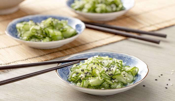 Fotografia em tons de verde em uma bancada de madeira com uma toalha de palha bege. Ao centro, um prato japonês branco com detalhes em azul e a salada de sunomono (pepinos fatiados bem finos) dentro, com um par de hashi apoiado.