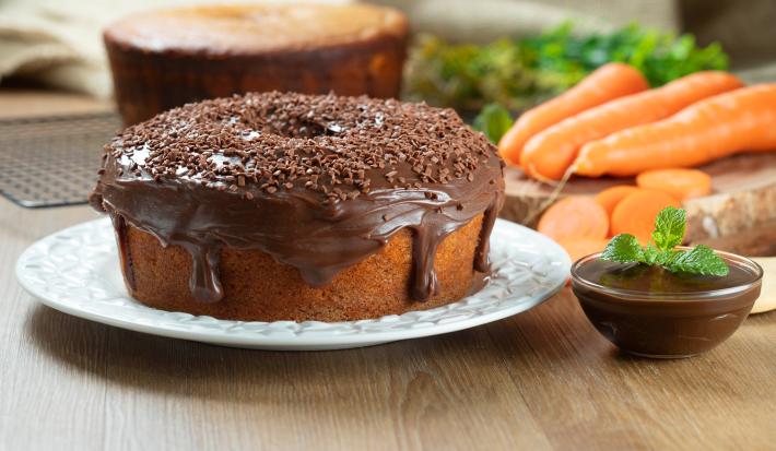 Fotografia de dois bolos de cenoura, um sem e o outro com cobertura de chocolate cremoso e granulado, apoiado em um prato branco. Ao lado tem mais cobertura em um recipiente pequeno de vidro, e ao fundo tem cenouras inteiras e alguns pedaços.