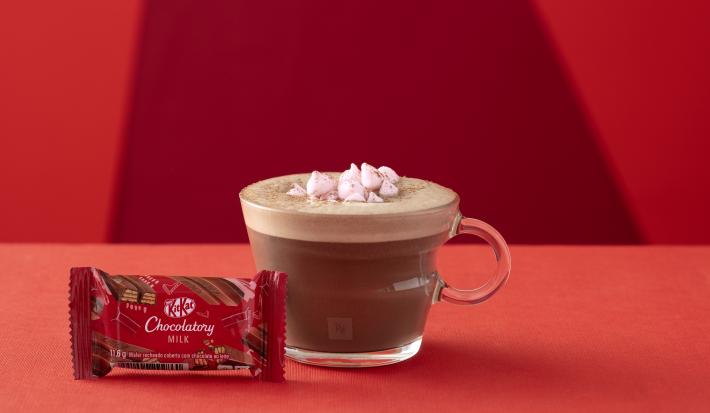 Foto da receita de Chocolate Quente. Observa-se um fundo vermelho com o chocolate quente em uma xícara de vidro transparente. Na frente, um kit kat fechado ao leite.