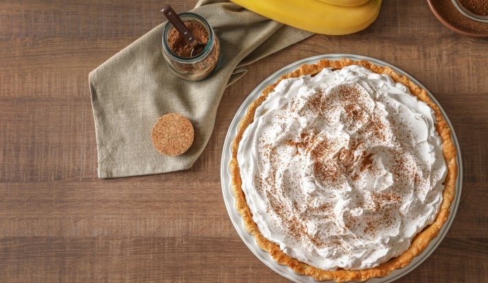 Fotografia vista de cima de uma torta banoffee inteira apoiada em um prato raso branco, sobre uma mesa de madeira.