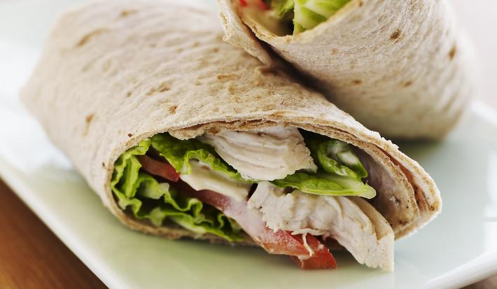 Fotografia de um wrap cortado ao meio, recheado de frango, alface, tomate e um molho. Os wraps estão em um prato branco quadrado, que está sobre uma mesa de madeira.