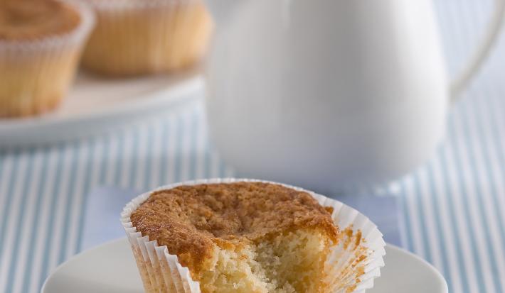 Fotografia em tons de azul em uma mesa de madeira com uma toalha listrada azul, um paninho azul, um pratinho de sobremesa branco com o muffin de banana com iogurte. Ao fundo, um bule branco com chá dentro.