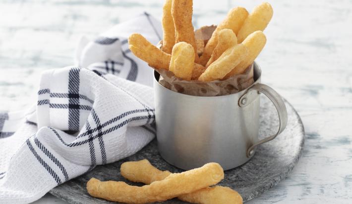 Imagem de uma caneca de alumínio com os palitos de biscoito de polvilho dourados. Ao lado, um paninho branco com detalhes preto e 2 biscoitos.