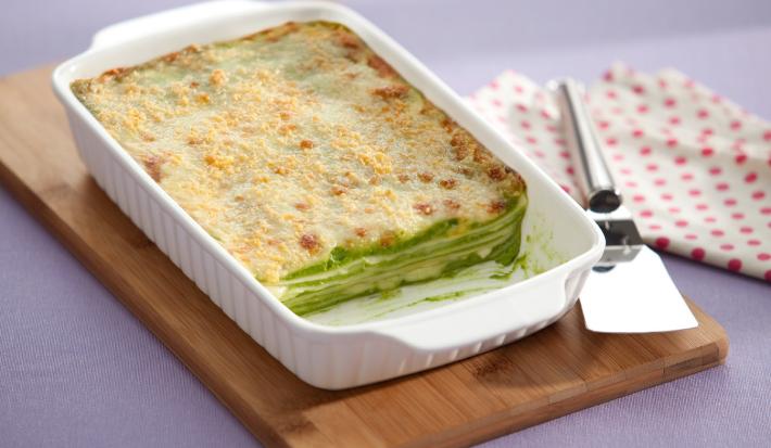 Fotografia em tons de branco e verde, com refratário branco contendo uma lasanha de espinafre gratinada, sobre uma tábua de cozinha com uma espátula de servir sobre um guardanapo branco com bolinhas vermelhas em uma bancada lilás.