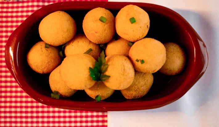 Fotografia vista de cima de bolinhas de queijo dentor de um recipiente oval marrom de vidro sobre um pano vermelho quadriculado com branco, o qual está apoaido em uma mesa branca.