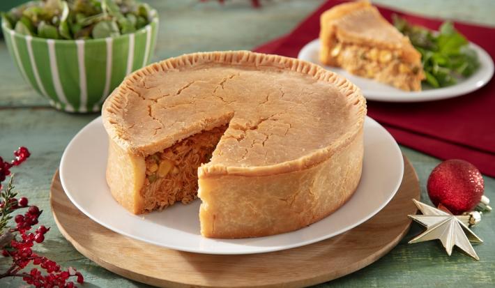 Foto da receita de empadão de frango sem glúten servida em um porção grande com uma fatia cortada ao fundo sobre um prato de porcelana branca. Ao fundo, há um paninho vermelho com decoração de natal