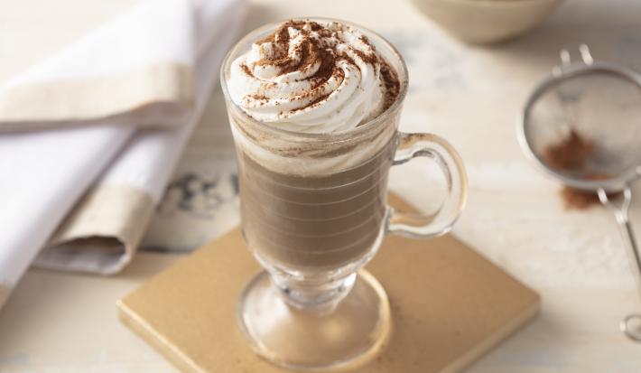 Fotografia em tons de marrom de uma bancada, ao centro uma placa quadrada bege e um copo transparente com alça e a bebida decorada com chantilly e chocolate em pó.